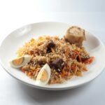 Home A savory dish of jollof rice served with meat, vegetables, and boiled eggs on a white plate.