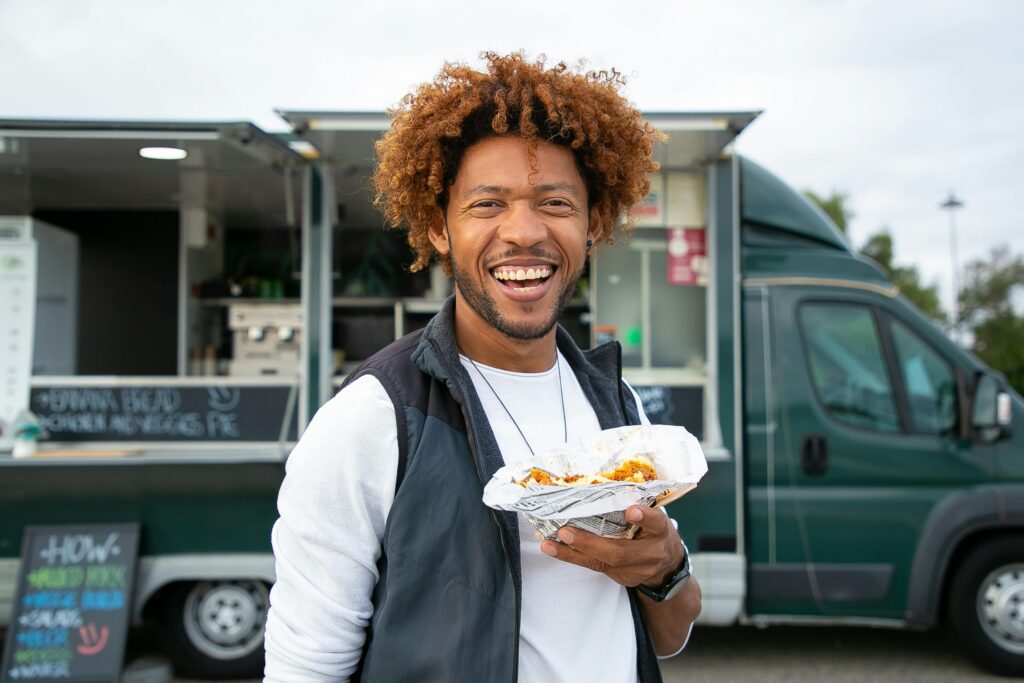 Smiling man holding street food near a food truck in a park setting.